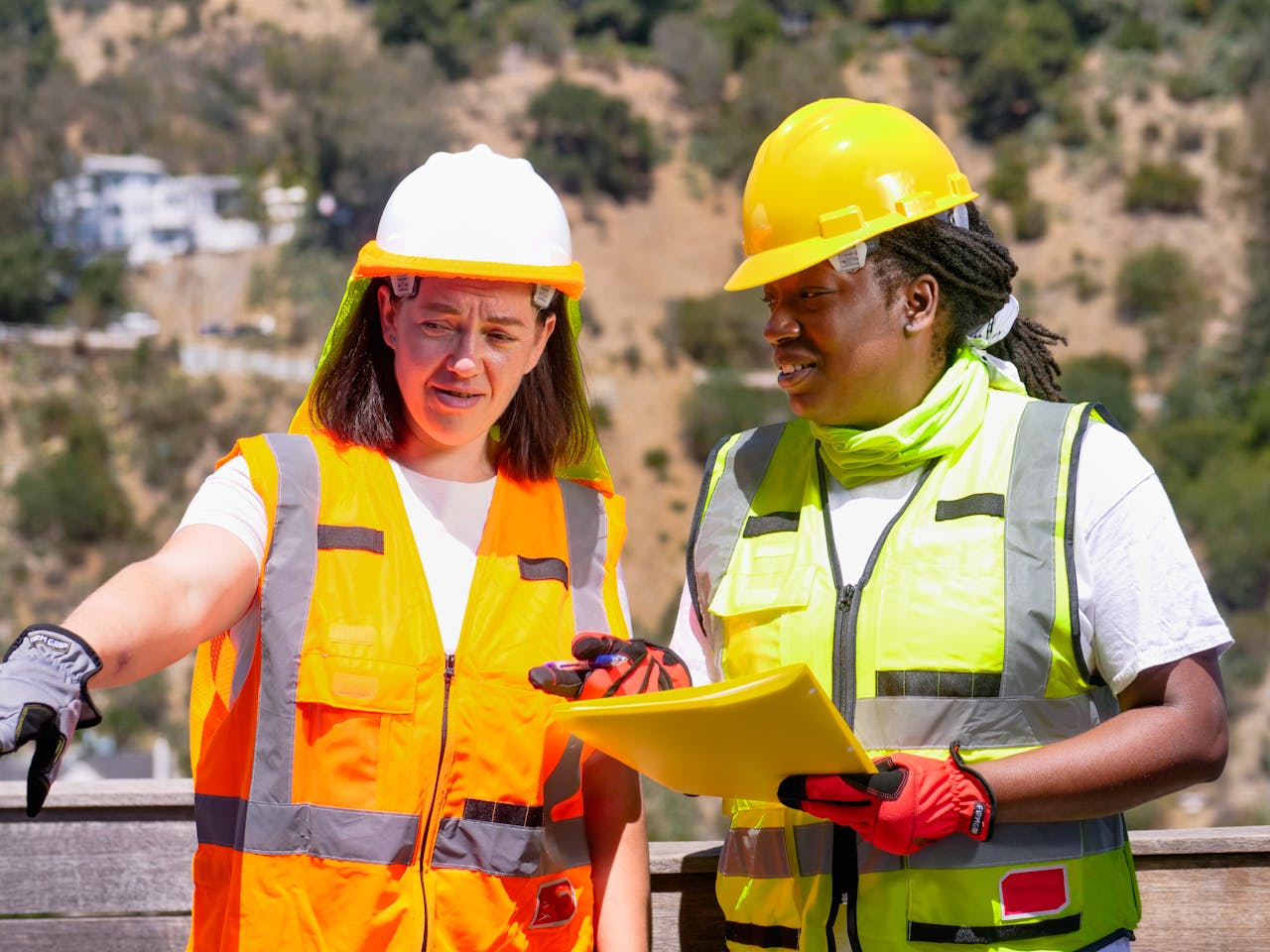 Two female engineers in safety gear discuss a project outdoors, focusing on plans.